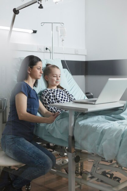 Giovane genitore e piccola figlia malata che navigano nelle pagine web sul computer portatile. Madre e figlia rilassate sedute nella stanza del reparto pazienti della clinica pediatrica mentre utilizzano un computer moderno.
