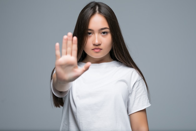 Giovane gesto asiatico serio di arresto di rappresentazione della donna con la sua palma mentre stando isolato sopra fondo grigio