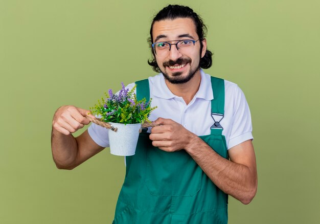 Giovane giardiniere barbuto uomo che indossa tuta che mostra pianta in vaso sorridente guardando davanti in piedi sulla parete verde chiaro