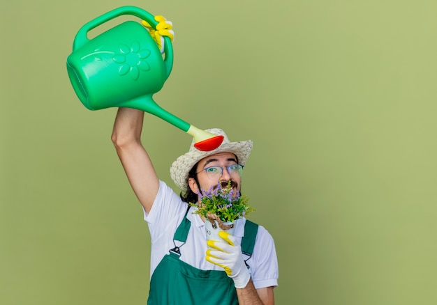 Giovane giardiniere barbuto uomo che indossa tuta e cappello azienda annaffiatoio e pianta in vaso impianto di irrigazione in piedi sopra il muro verde chiaro