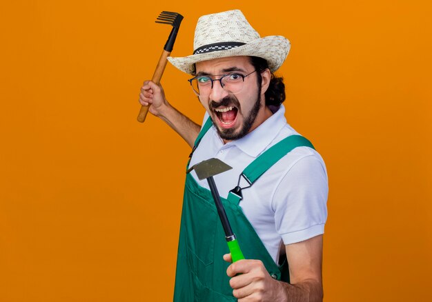 Giovane giardiniere barbuto uomo che indossa tuta e cappello azienda mattock e mini rastrello guardando davanti gridando con faccia arrabbiata in piedi sopra la parete arancione