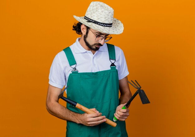 Giovane giardiniere barbuto uomo che indossa tuta e cappello azienda mattock e mini rastrello guardandoli confusi in piedi sopra la parete arancione