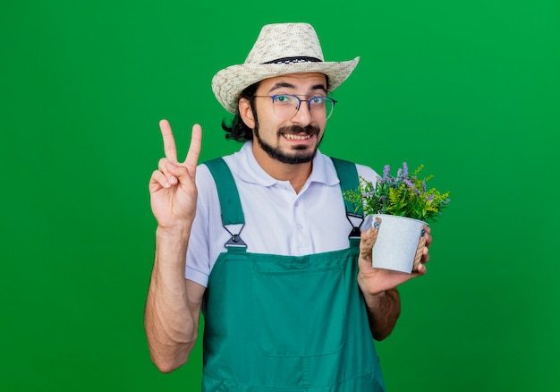 Giovane giardiniere barbuto uomo che indossa tuta e cappello azienda pianta in vaso che mostra v-segno