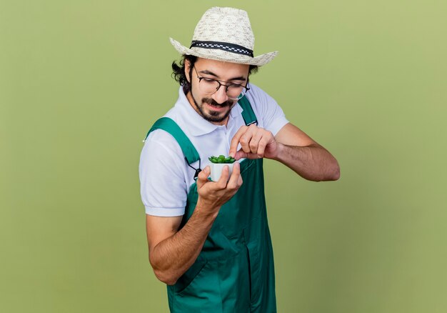 Giovane giardiniere barbuto uomo che indossa tuta e cappello che mostra pianta in vaso guardandolo sorridente in piedi sopra il muro verde chiaro