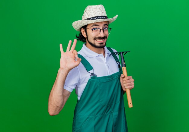 Giovane giardiniere barbuto uomo che indossa tuta e cappello che tiene mini rastrello sorridente che mostra segno giusto