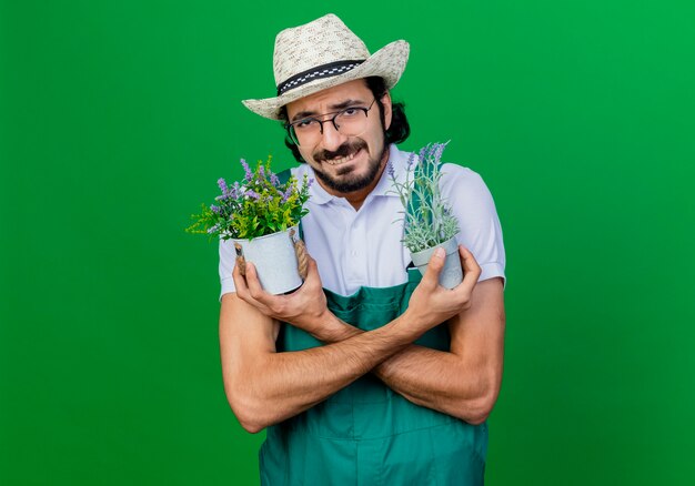 Giovane giardiniere barbuto uomo che indossa tuta e cappello che tiene piante in vaso preoccupato di essere scontento