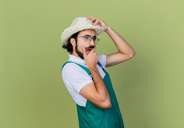 Giovane giardiniere barbuto uomo che indossa tuta e cappello guardando da parte di essere sorpreso e stupito in piedi sopra la parete verde chiaro