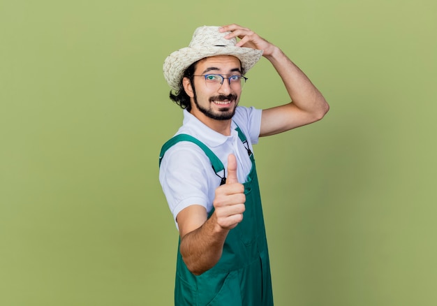 Giovane giardiniere barbuto uomo che indossa tuta e cappello guardando la parte anteriore sorridente che mostra i pollici in su in piedi sopra il muro verde chiaro