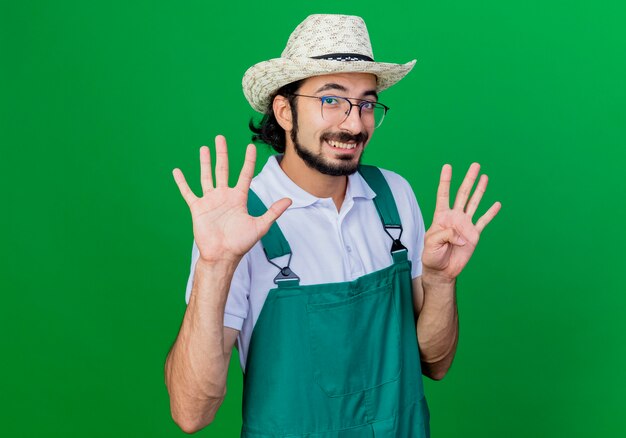 Giovane giardiniere barbuto uomo che indossa tuta e cappello sorridente che mostra il numero nove