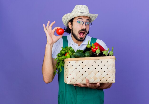 Giovane giardiniere barbuto uomo che indossa tuta e cappello tenendo la cassa piena di verdure che mostra il pomodoro fresco di essere sorpreso in piedi sopra la parete blu