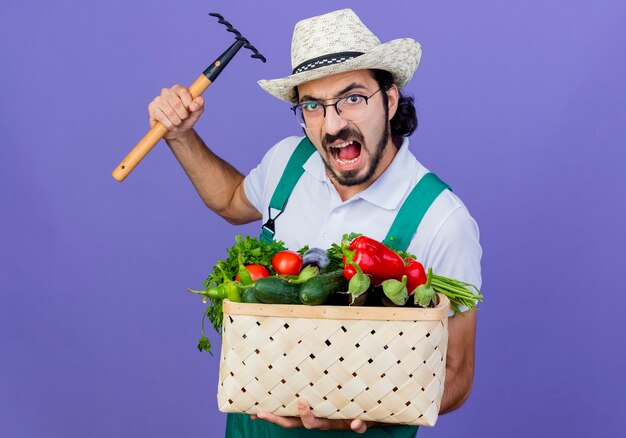 Giovane giardiniere barbuto uomo che indossa tuta e cappello tenendo la cassa piena di verdure facendo oscillare un mini rastrello con la faccia arrabbiata in piedi sopra la parete blu