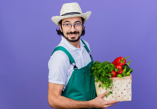 Giovane giardiniere barbuto uomo che indossa tuta e cappello tenendo la cassa piena di verdure guardando la parte anteriore sorridente allegramente in piedi sopra la parete blu