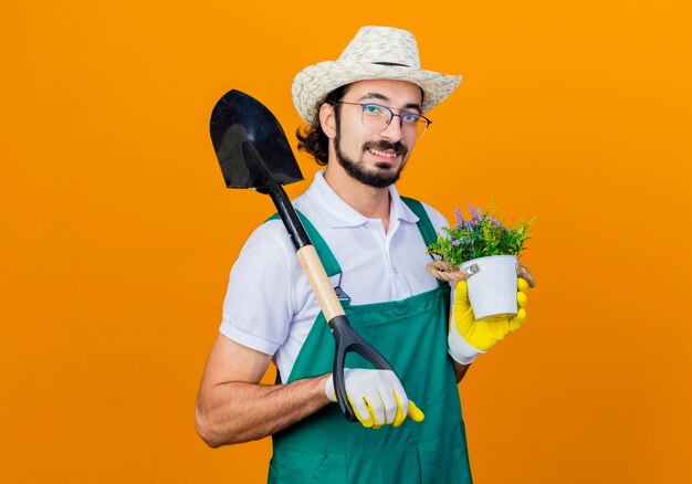 Giovane giardiniere barbuto uomo che indossa tuta e cappello tenendo la pala e pianta in vaso guardando la parte anteriore sorridente con la faccia felice in piedi sopra la parete arancione