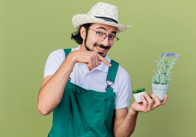 Giovane giardiniere barbuto uomo che indossa tuta e cappello tenendo la pianta in vaso puntando con il dito indice su di esso sorridendo allegramente in piedi sopra il muro verde chiaro