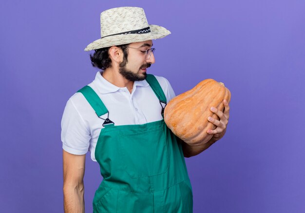 Giovane giardiniere barbuto uomo che indossa tuta e cappello tenendo la zucca guardando sorridente in piedi sopra la parete blu