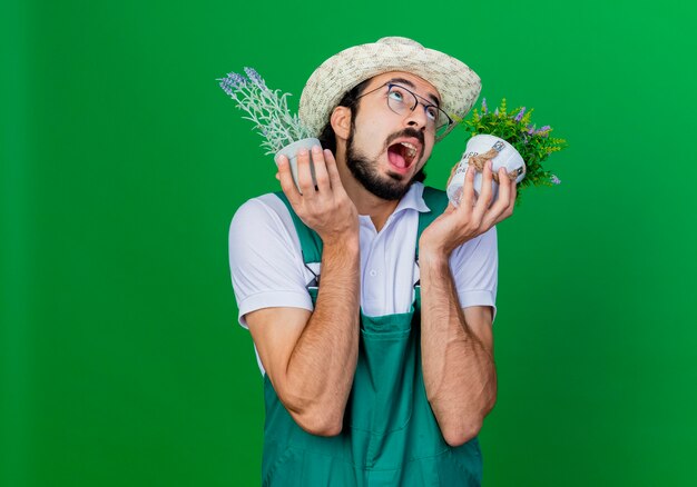 Giovane giardiniere barbuto uomo che indossa tuta e cappello tenendo le piante in vaso cercando felice ed emozionato