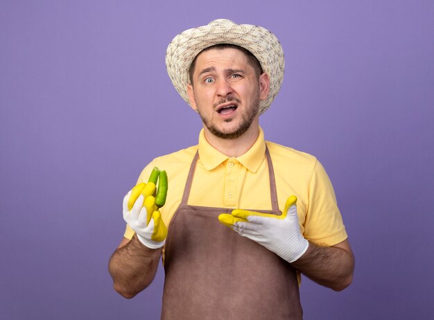 Giovane giardiniere che indossa tuta e cappello in guanti da lavoro che tengono peperoncino verde rotto presentandolo con il braccio che guarda davanti essendo confuso e dispiaciuto in piedi sopra il muro viola