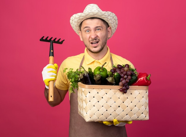 Giovane giardiniere che indossa tuta e cappello in guanti da lavoro tenendo la cassa piena di verdure che mostra mini rastrello guardando davanti confuso in piedi sopra il muro rosa