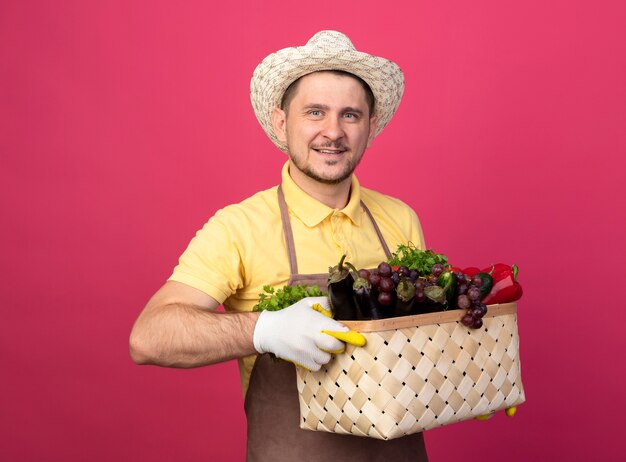 Giovane giardiniere che indossa tuta e cappello in guanti da lavoro tenendo la cassa piena di verdure guardando davanti sorridente felice e positivo in piedi sopra il muro rosa