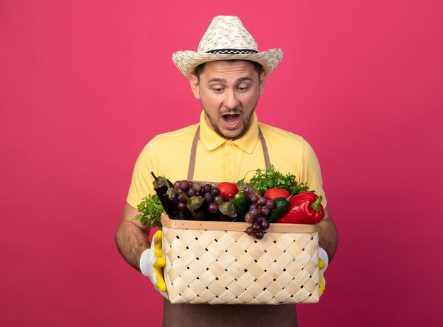 Giovane giardiniere che indossa tuta e cappello in guanti da lavoro tenendo la cassa piena di verdure guardando stupito e sorpreso in piedi sopra il muro rosa