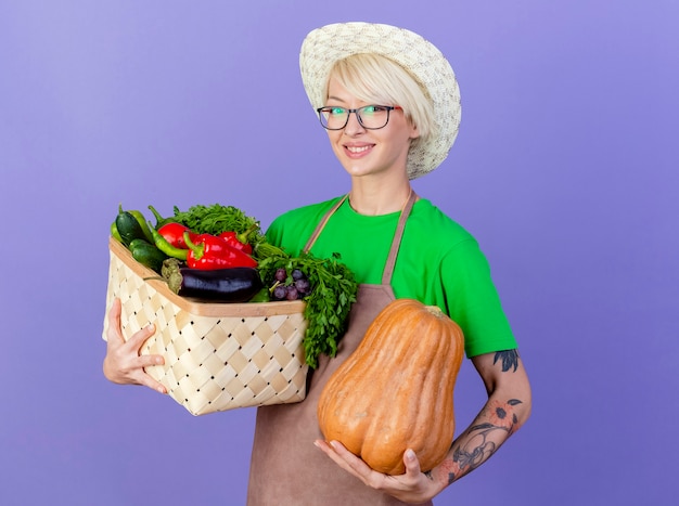 Giovane giardiniere donna con i capelli corti in grembiule e cappello azienda cassa piena di verdure e zucca lookign in telecamera sorridendo allegramente in piedi su sfondo blu