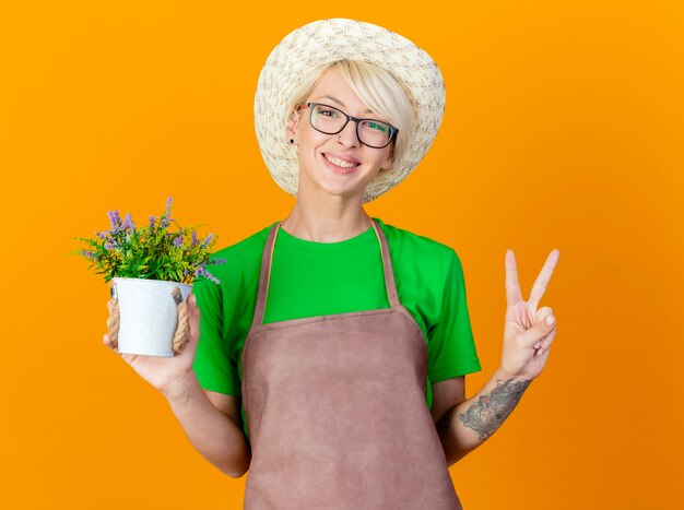 Giovane giardiniere donna con i capelli corti in grembiule e cappello azienda pianta in vaso guardando la telecamera sorridendo con la faccia felice che mostra v-segno in piedi su sfondo arancione