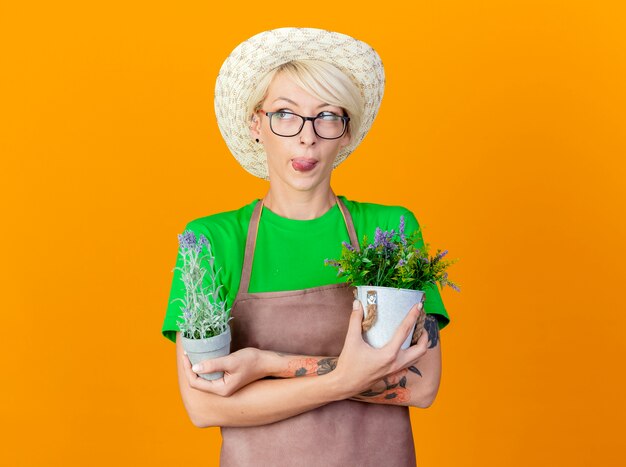 Giovane giardiniere donna con i capelli corti in grembiule e cappello azienda piante in vaso lookign da parte spuntavano lingua in piedi su sfondo arancione