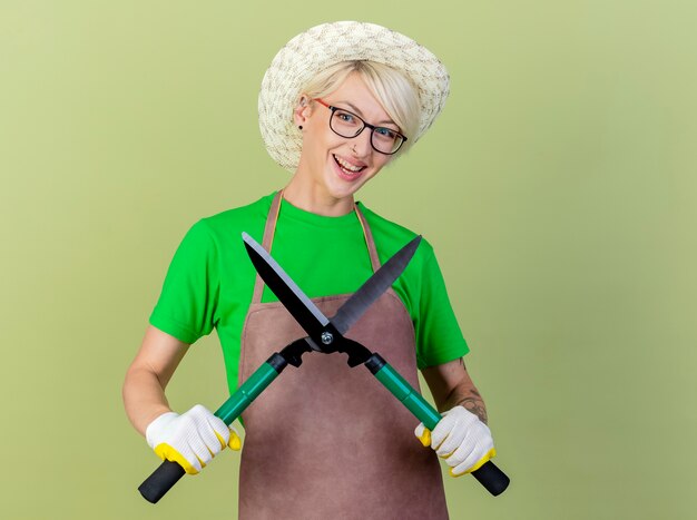 Giovane giardiniere donna con i capelli corti in grembiule e cappello azienda tagliasiepi guardando la telecamera sorridendo allegramente in piedi su sfondo chiaro