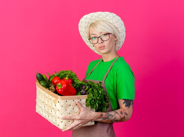 Giovane giardiniere donna con i capelli corti in grembiule e cappello tenendo la cassa piena di verdure