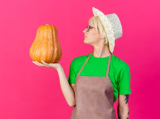 Giovane giardiniere donna con i capelli corti in grembiule e cappello tenendo la zucca