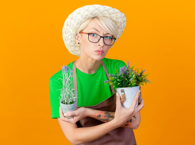 Giovane giardiniere donna con i capelli corti in grembiule e cappello tenendo le piante in vaso guardando la telecamera confusa in piedi su sfondo arancione