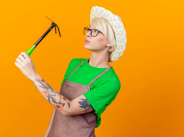 Giovane giardiniere donna con i capelli corti in grembiule e cappello tenendo mini rastrello in piedi su sfondo arancione