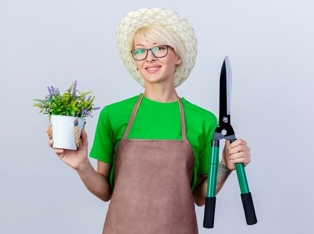 Giovane giardiniere donna con i capelli corti in grembiule e cappello tenendo tagliasiepi e pianta in vaso guardando la telecamera sorridente con la faccia felice in piedi su sfondo bianco