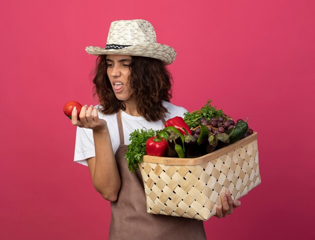 Giovane giardiniere femminile dispiaciuto in cappello da giardinaggio da portare uniforme che tiene il cestino della verdura e che esamina il pomodoro in sua mano isolata sul colore rosa