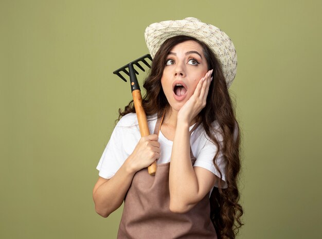 Giovane giardiniere femminile scioccato in uniforme che indossa il cappello da giardinaggio mette la mano sul viso e tiene il rastrello guardando il lato isolato sulla parete verde oliva