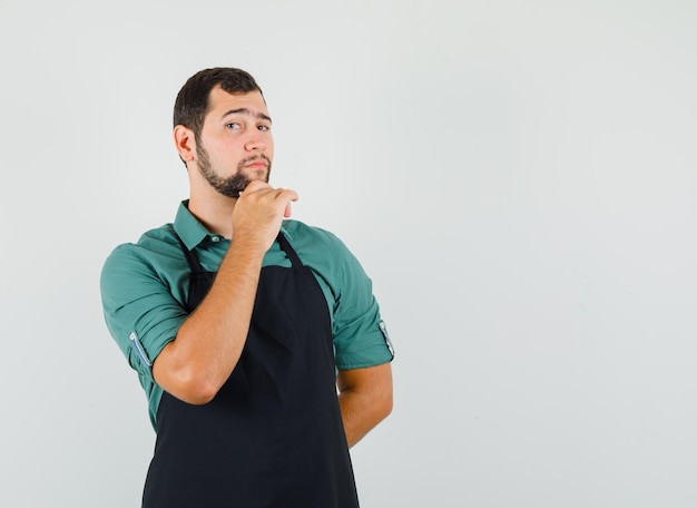 Giovane giardiniere in t-shirt, grembiule appoggiato al mento e guardando premuroso, vista frontale. spazio per il testo