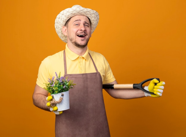 Giovane giardiniere in tuta e cappello tenendo la pala e la pianta in vaso guardando la parte anteriore sorridente con la faccia felice in piedi sopra la parete arancione