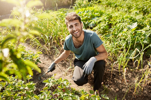 Giovane giardiniere maschio barbuto attraente allegro in maglietta blu e pantaloni sportivi neri sorridente, lavorando in giardino, piantando germogli con la pala.
