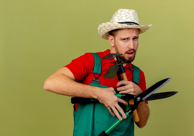 Giovane giardiniere slavo bello dispiaciuto in uniforme e cappello che tengono gli strumenti del giardiniere che sembrano isolati