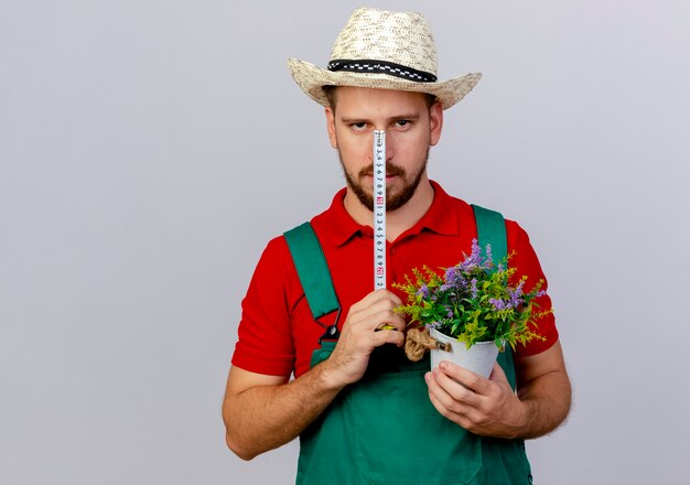 Giovane giardiniere slavo bello in uniforme e metro a nastro della tenuta del cappello davanti al fronte che osserva con il vaso di fiori in un'altra mano isolata