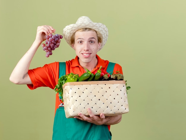 Giovane giardiniere uomo che indossa tuta e cappello azienda cassa piena di verdure e grappolo d'uva guardando la fotocamera con il sorriso sul viso in piedi su sfondo chiaro