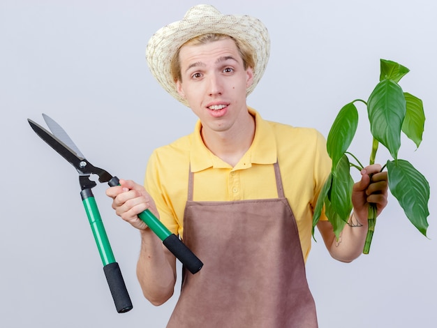 Giovane giardiniere uomo che indossa tuta e cappello azienda pianta e tagliasiepi guardando la telecamera sorridente con la faccia felice in piedi su sfondo bianco
