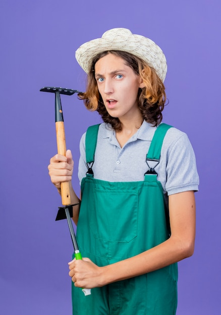 Giovane giardiniere uomo che indossa tuta e cappello che tiene mini rastrello con faccia seria