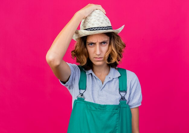 Giovane giardiniere uomo che indossa tuta e cappello con la faccia seria che tocca il suo cappello