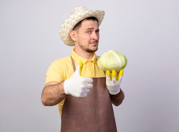 Giovane giardiniere uomo che indossa tuta e cappello in guanti da lavoro tenendo il cavolo lookign alla telecamera che mostra i pollici in su