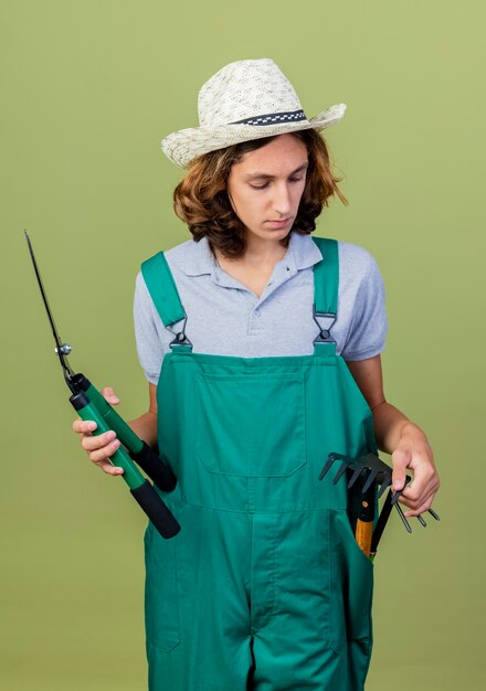 Giovane giardiniere uomo che indossa tuta e cappello tenendo attrezzature da giardinaggio