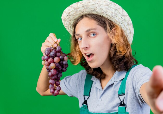 Giovane giardiniere uomo che indossa tuta e cappello tenendo il mazzo di uva sorridente