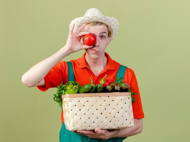 Giovane giardiniere uomo che indossa tuta e cappello tenendo la cassa piena di verdure che coprono gli occhi con il pomodoro sorridente in piedi su sfondo chiaro