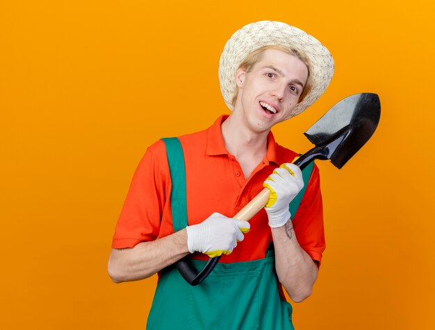 Giovane giardiniere uomo che indossa tuta e cappello tenendo la pala sorridente