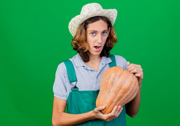 Giovane giardiniere uomo che indossa tuta e cappello tenendo la zucca sorridente